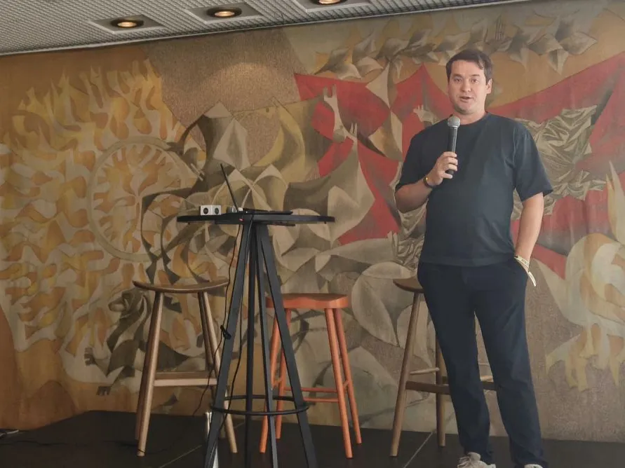 A speaker holding a microphone addresses the room from the SoAlive stage in Sofia, in front of a large painted mural and three wooden bar stools