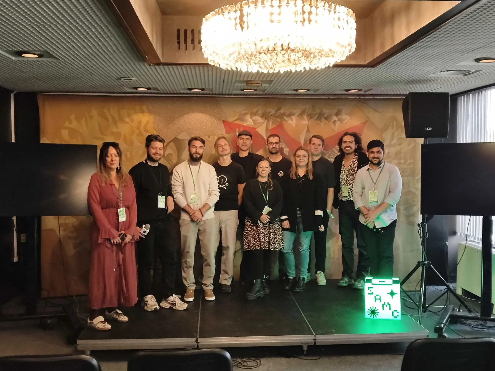 Eleven people from the SoAlive Startup Showcase 2026 line up on a low stage in Sofia, under a crystal chandelier and in front of a painted mural, with a glowing SAMC sign at their feet